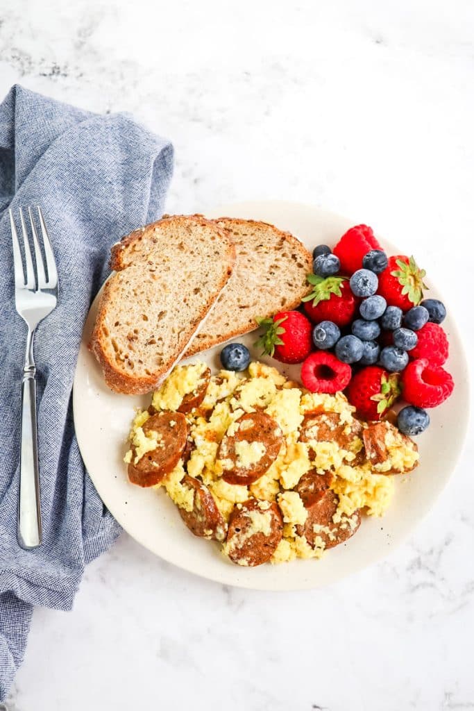 Egg and sausage scramble with fresh berries and toast on a plate with fork and napkin on the side.