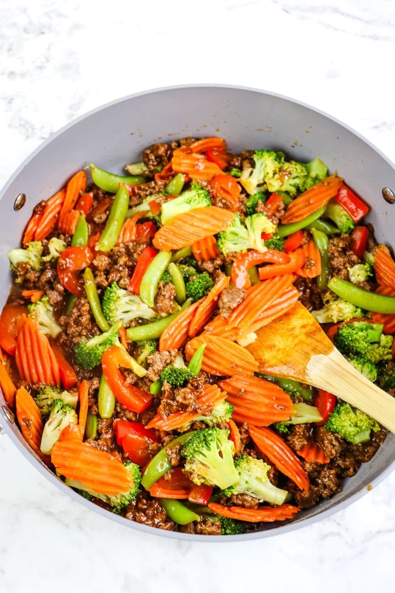Ground beef stir fry with steamed vegetables stirred together with a wooden spoon.