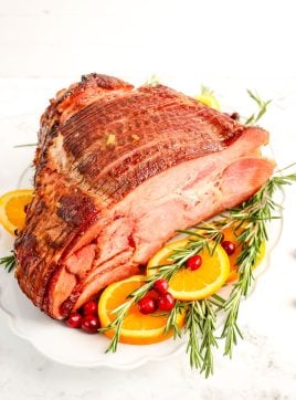 Orange glazed ham on a serving platter with rosemary, cranberry and orange slices for garnish.