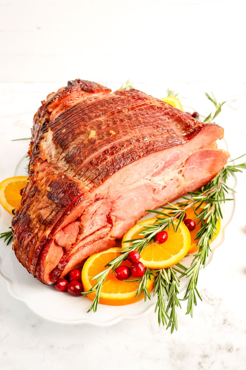 Orange glazed ham on a serving platter with rosemary, cranberry and orange slices for garnish.