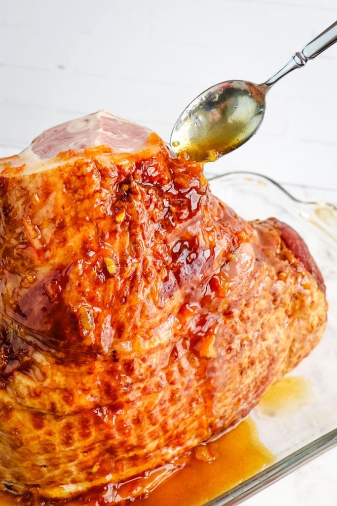 Orange glaze being spooned over a spiral ham in a baking dish.