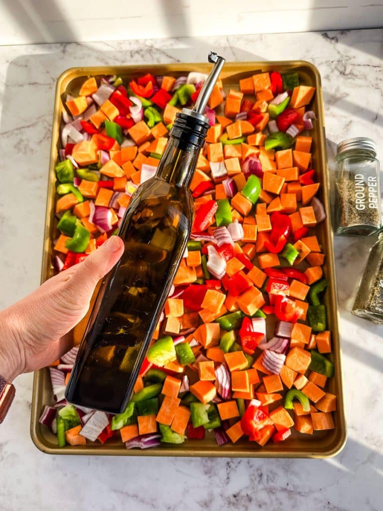 Chopped sweet potatoes, bell peppers and onions on a sheet pan being drizzled with olive oil.