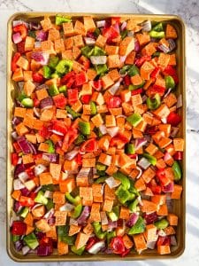 Cubed sweet potatoes, red and green bell peppers and red onion all on a sheet pan ready to be baked.