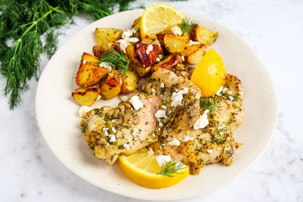 Greek chicken thighs and lemon potatoes on a plate, garnished with lemon slices and feta cheese crumbles.